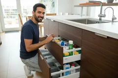 Homem a organizar gavetas de cozinha moderna junto ao lavatório com produtos e utensílios.
