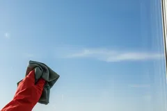 Mão com luva vermelha limpa janela de vidro com pano sob céu azul em Portugal.