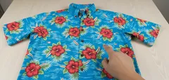 Camisa havaiana azul com flores vermelhas sendo apontada por uma mão, sobre uma mesa.