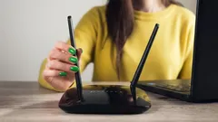 Mulher ajusta antena de router Wi-Fi ao lado de computador portátil em mesa de madeira.
