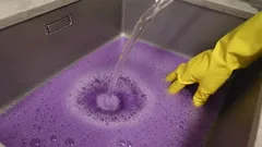 Líquido roxo em pia de inox com mão de luva amarela e água a correr.